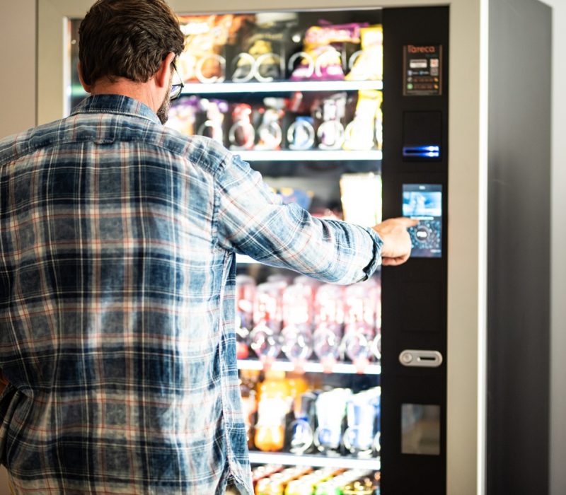 Back view of adult mature man choosing snacks and drinks on 24h free automated distributor - people buying food on airport gate - travel lifestyle and junk nutrition unhealthy lifestyle